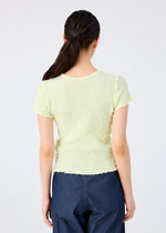 A woman with long brown hair in a low ponytail is seen from the back wearing the CAULIFLOWER COLOR top by me ISSEY MIYAKE and dark blue jeans, set against a plain light background.