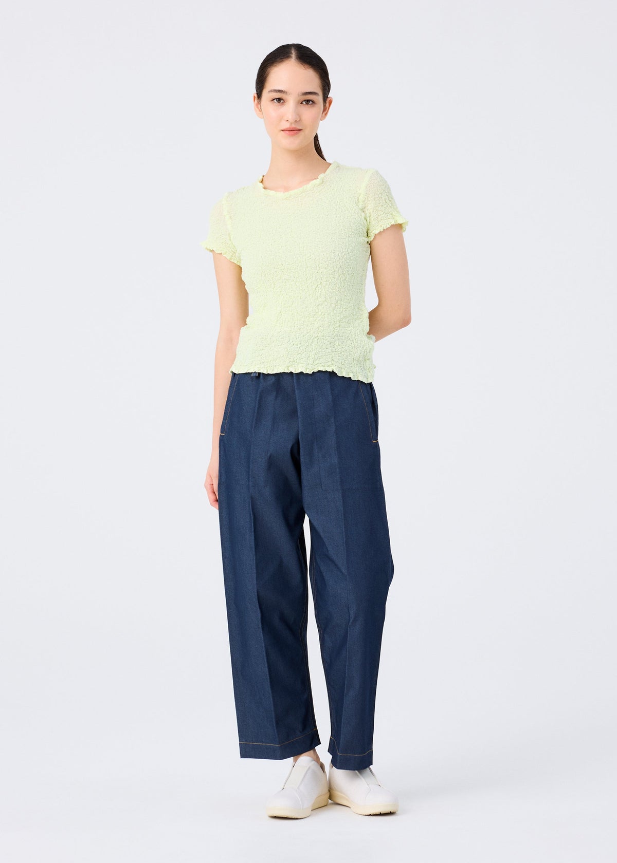 A woman poses against a white background, wearing the CAULIFLOWER COLOR light yellow round-neck top by me ISSEY MIYAKE, paired with loose dark blue jeans and white shoes. Her brown hair is tied back, hands behind her. view 7 of 7