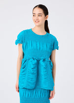 A woman with long dark hair tied back wears the SHRINK STRIPE short-sleeve dress by me ISSEY MIYAKE, featuring bright blue cotton-blend fabric with gathered details. She faces right and smiles against a plain light background.