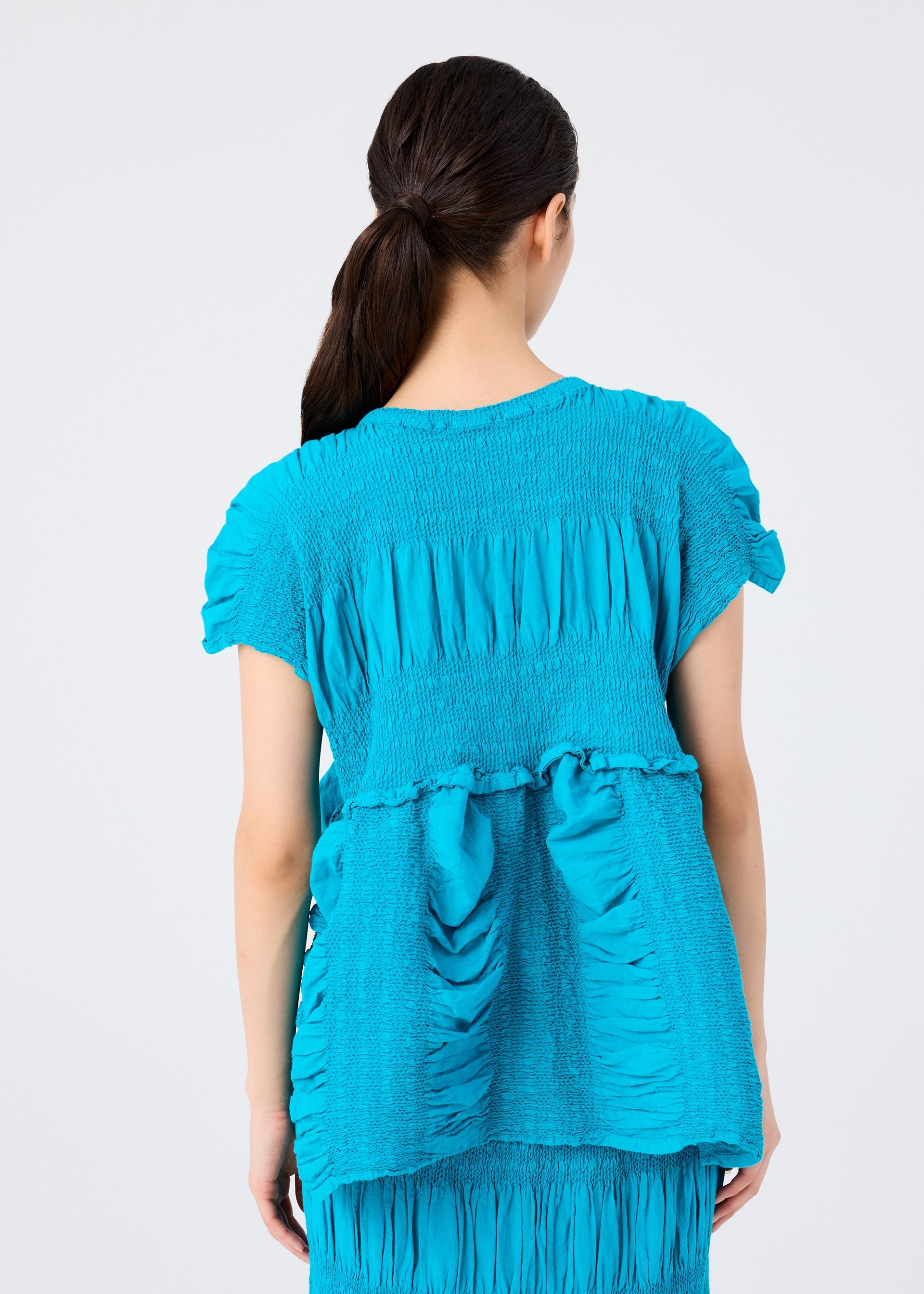 Zoomed view A woman with long brown hair in a ponytail is seen from behind wearing the me ISSEY MIYAKE SHRINK STRIPE top in bright blue, which features textured, ruched cotton-blend fabric on the back.