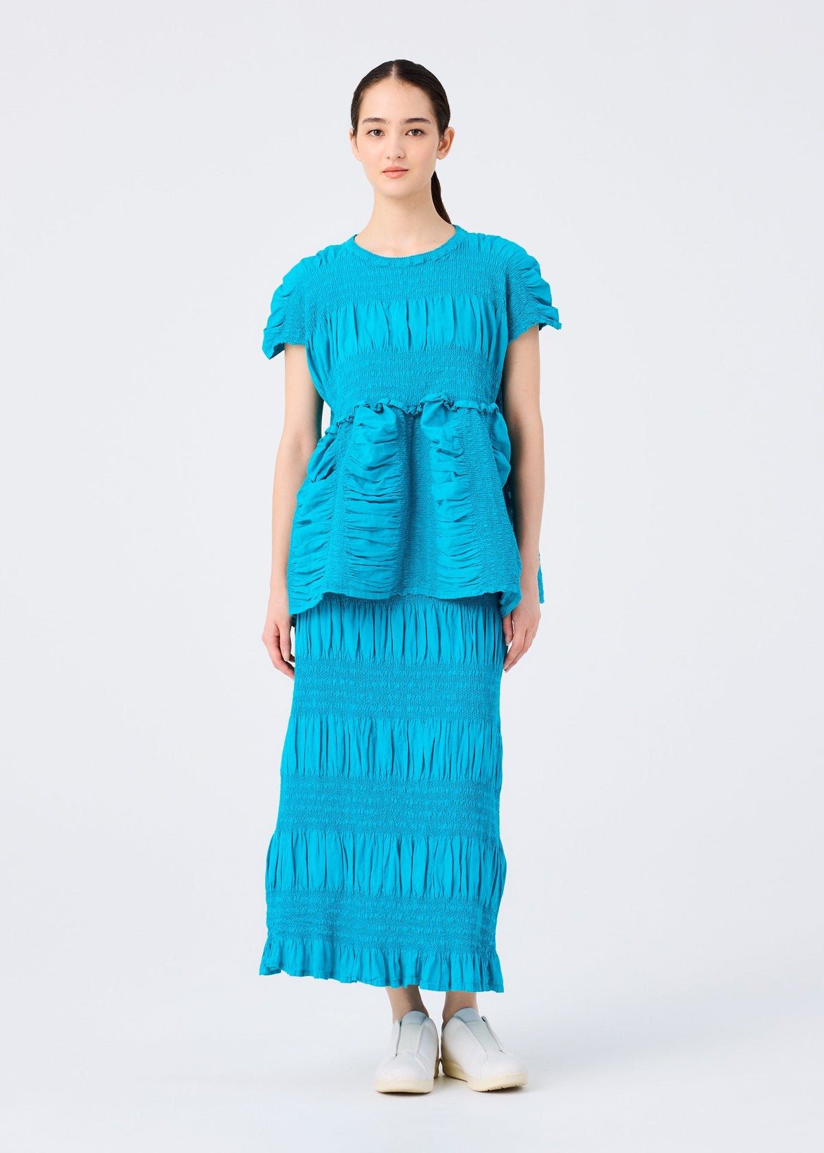 A woman faces forward, wearing the SHRINK STRIPE cotton-blend peplum top and long matching skirt by me ISSEY MIYAKE. She has straight dark hair pulled back, white slip-on shoes, and stands against a plain white background. view 7 of 7