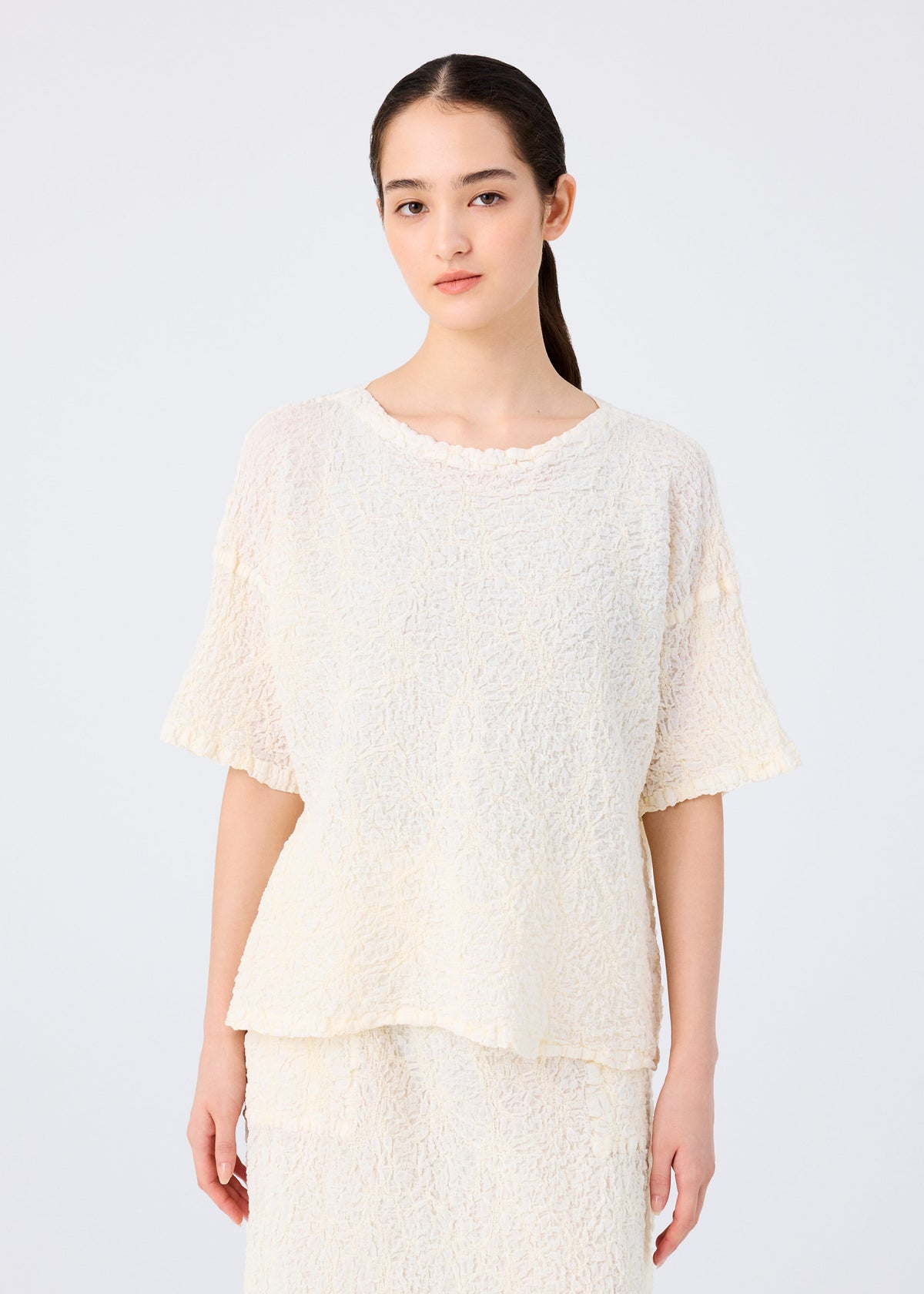 A woman with long dark hair tied back wears the SURPRISE BOX PRINT CAULIFLOWER set by me ISSEY MIYAKE, featuring a loose, textured cream cotton short-sleeved top and matching skirt, against a plain light background. view 5 of 7