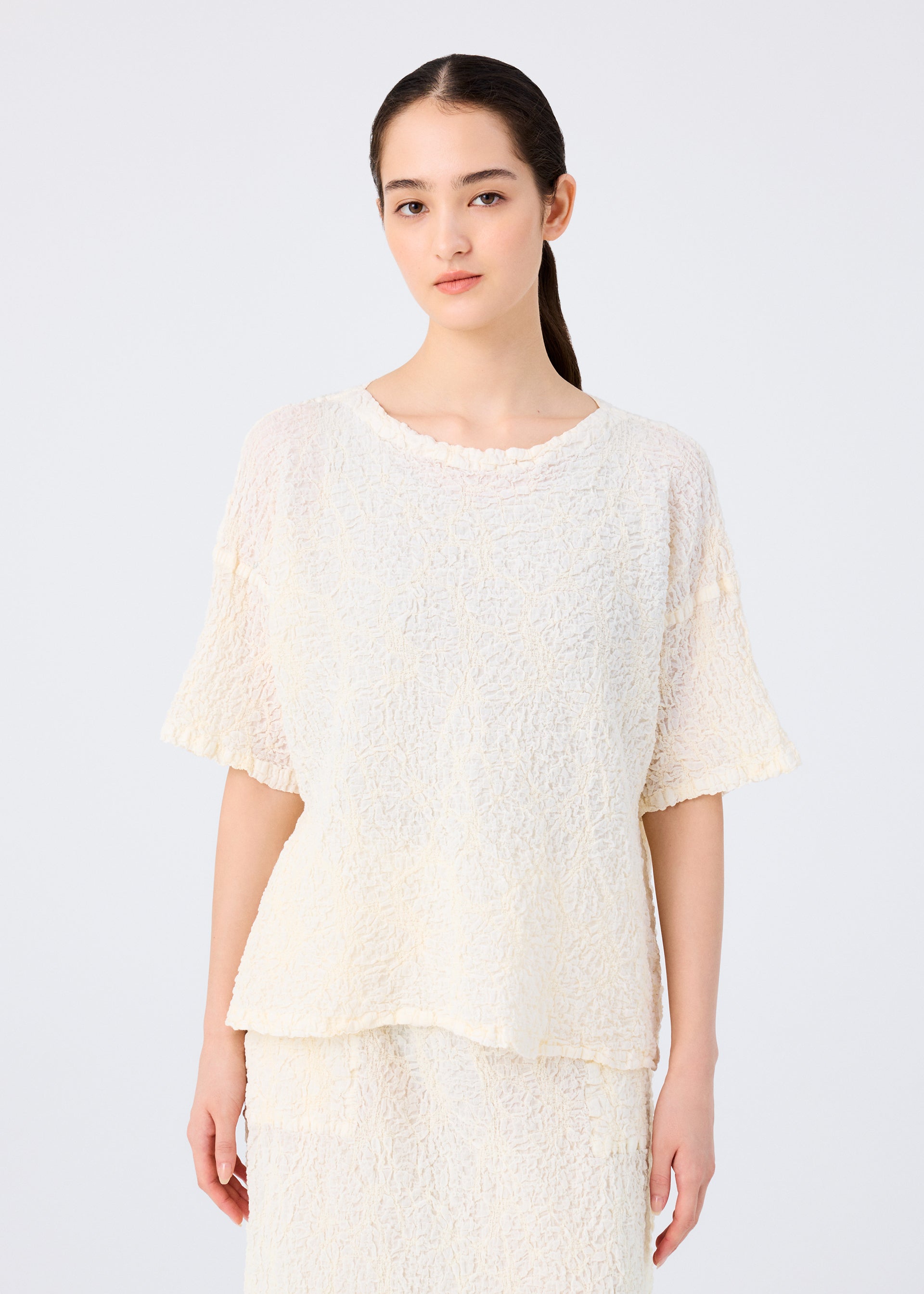 Zoomed view A woman with long dark hair tied back wears the SURPRISE BOX PRINT CAULIFLOWER set by me ISSEY MIYAKE, featuring a loose, textured cream cotton short-sleeved top and matching skirt, against a plain light background.