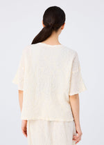 A woman with long dark hair in a low ponytail is seen from behind, wearing the me ISSEY MIYAKE SURPRISE BOX PRINT CAULIFLOWER set against a plain light background.