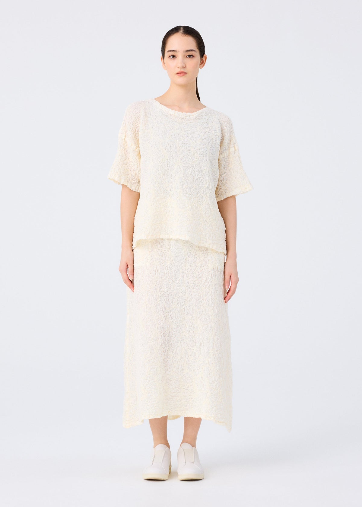 A woman stands facing forward in a loose-fitting SURPRISE BOX PRINT CAULIFLOWER top and matching midi skirt by me ISSEY MIYAKE, both in textured cream fabric. Her hair is pulled back, with white slip-on shoes against a plain white background. view 7 of 7