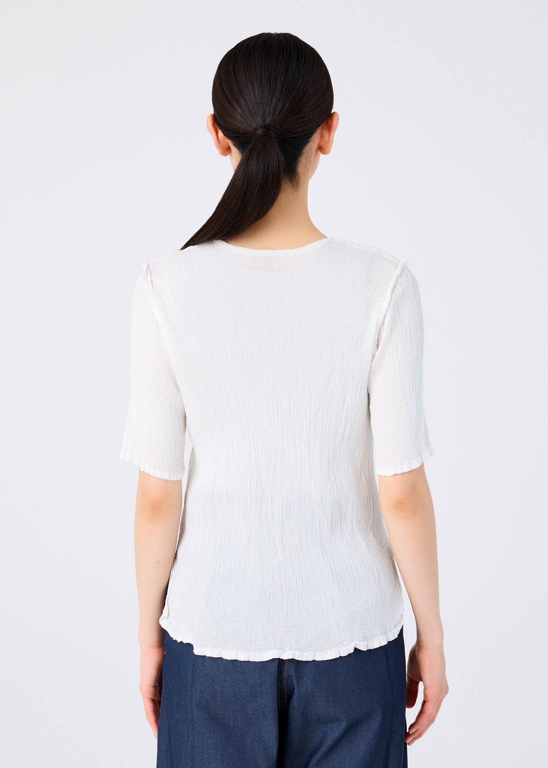 Zoomed view A person with long dark hair in a low ponytail is seen from behind, wearing the SEE-THROUGH CREPE COLOR white top by me ISSEY MIYAKE and dark blue pants, standing against a plain light background.