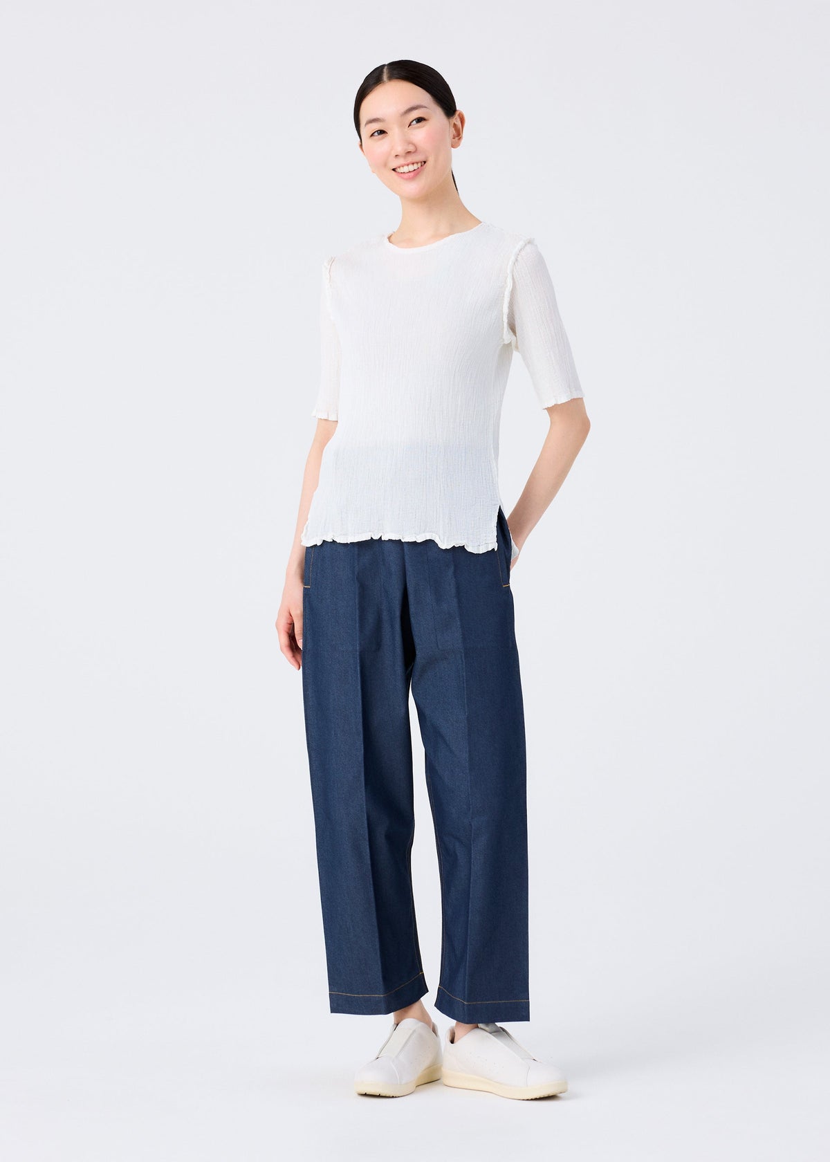 A person stands smiling against a plain white backdrop, wearing the SEE-THROUGH CREPE COLOR top by me ISSEY MIYAKE, loose dark blue pants, and white slip-on shoes, hands in pockets. view 8 of 8