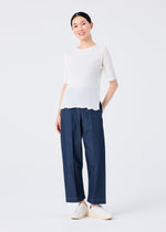A person stands smiling against a plain white backdrop, wearing the SEE-THROUGH CREPE COLOR top by me ISSEY MIYAKE, loose dark blue pants, and white slip-on shoes, hands in pockets.