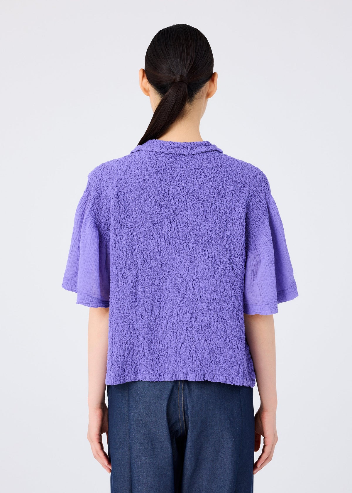A person with long dark hair in a low ponytail is shown from behind, wearing the SHEER CAULIFLOWER short-sleeved top by me ISSEY MIYAKE and dark blue pants against a plain white background. view 6 of 7