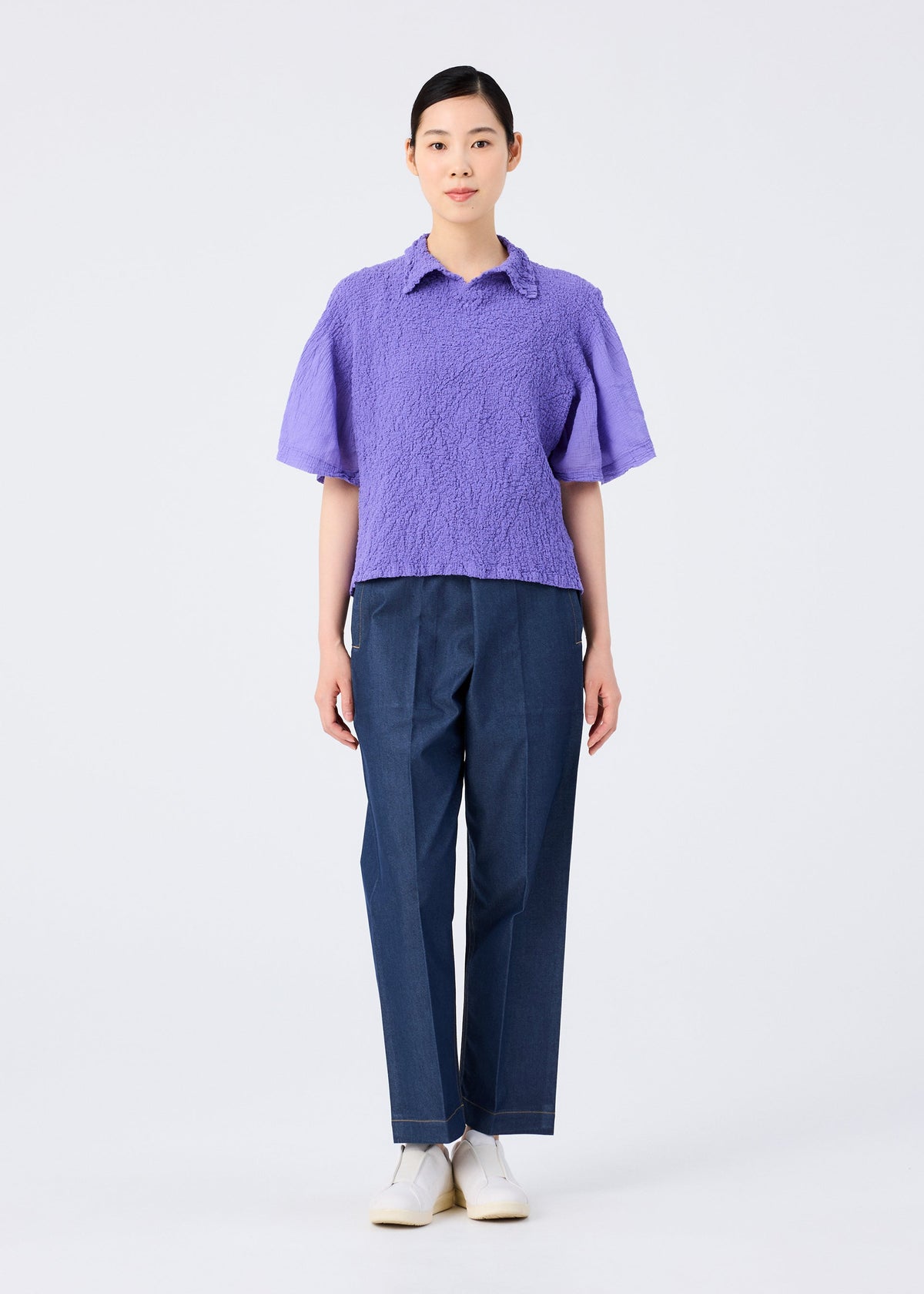 A person stands facing forward in a me ISSEY MIYAKE SHEER CAULIFLOWER lavender short-sleeved top, dark blue trousers, and white shoes against a plain white background. view 7 of 7