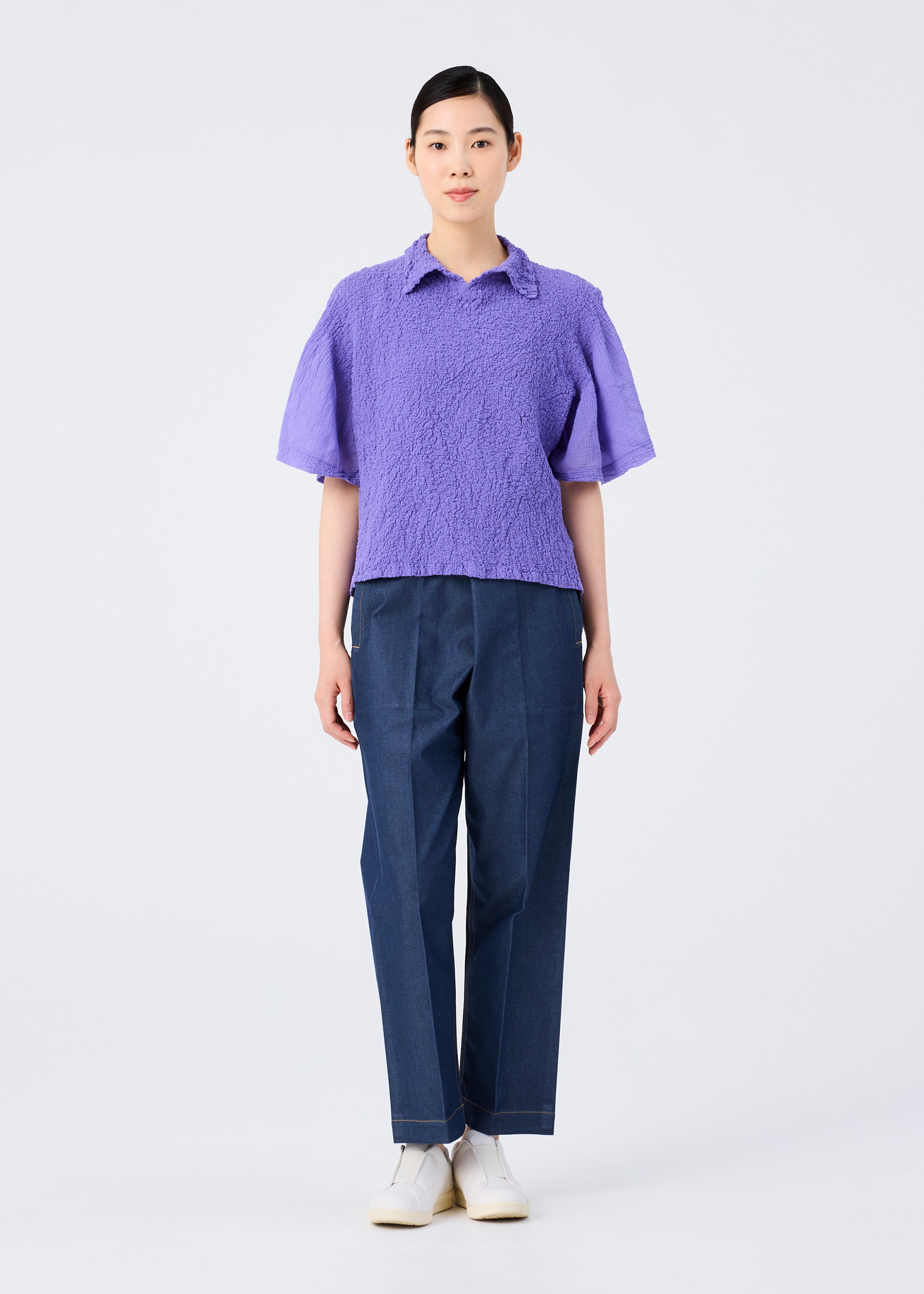 Zoomed view A person stands facing forward in a me ISSEY MIYAKE SHEER CAULIFLOWER lavender short-sleeved top, dark blue trousers, and white shoes against a plain white background.