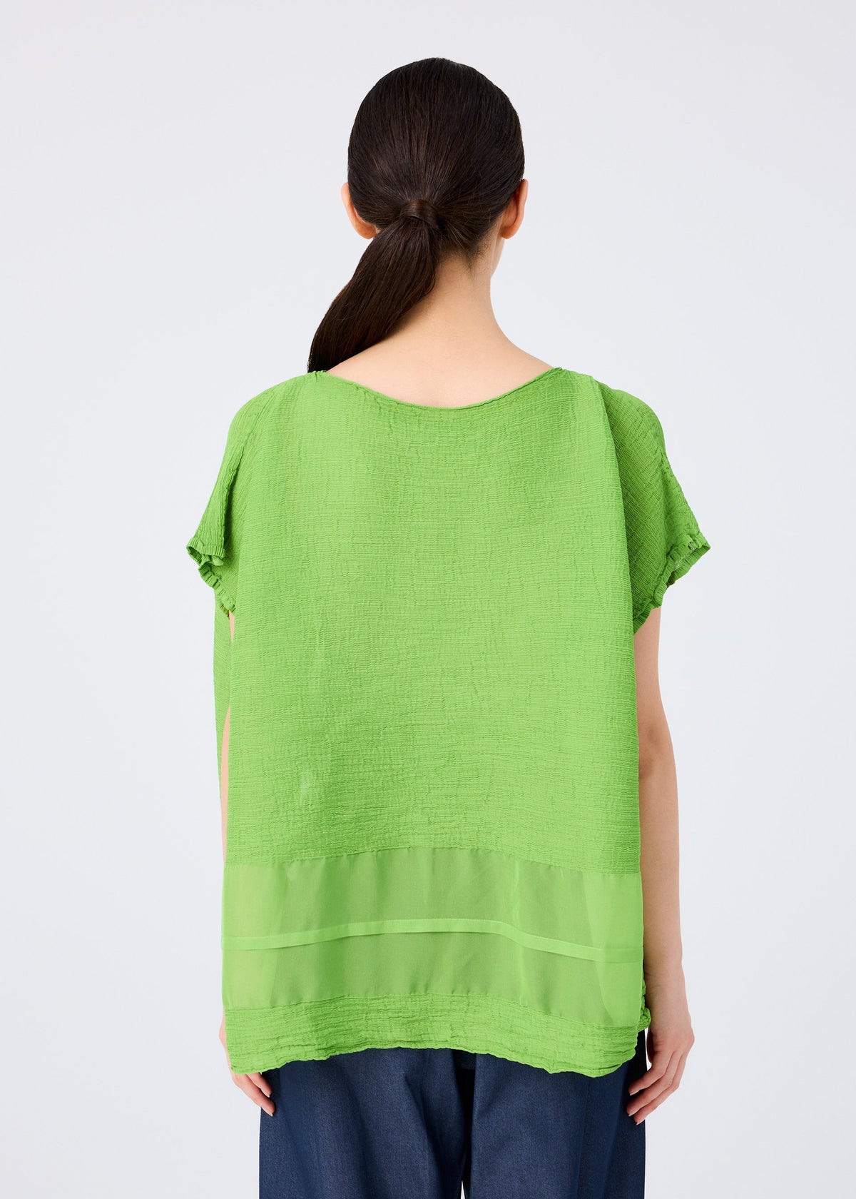 A person with long dark hair in a low ponytail is shown from the back wearing me ISSEY MIYAKE’s SHEER SEE-THROUGH CREPE textured green top with a sheer panel near the hem and dark pants, set against a plain light background. view 6 of 7