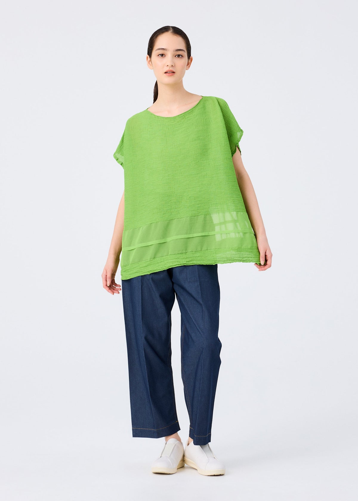 A woman faces forward in a bright green SHEER SEE-THROUGH CREPE top by me ISSEY MIYAKE with sheer details, paired with wide-leg dark blue pants and white shoes. Her dark hair is pulled back, set against a plain light background. view 7 of 7