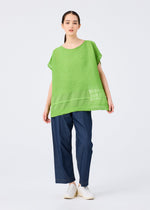 A woman faces forward in a bright green SHEER SEE-THROUGH CREPE top by me ISSEY MIYAKE with sheer details, paired with wide-leg dark blue pants and white shoes. Her dark hair is pulled back, set against a plain light background.