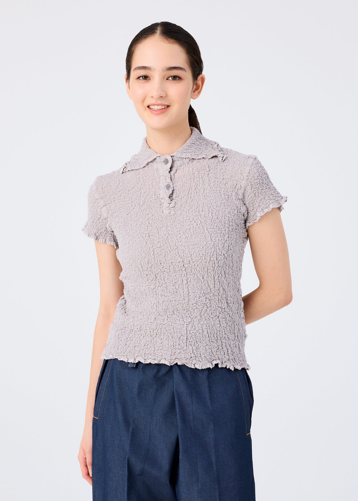 A young woman with long dark hair in a ponytail smiles at the camera, wearing the CAULIFLOWER COLOR top by me ISSEY MIYAKE in light gray, paired with dark blue pants, against a plain light background. view 5 of 7