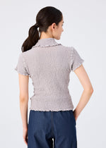 A woman with dark hair in a low ponytail wears the stretchy short-sleeved CAULIFLOWER COLOR top by me ISSEY MIYAKE and dark blue pants, shown from behind against a plain light background.