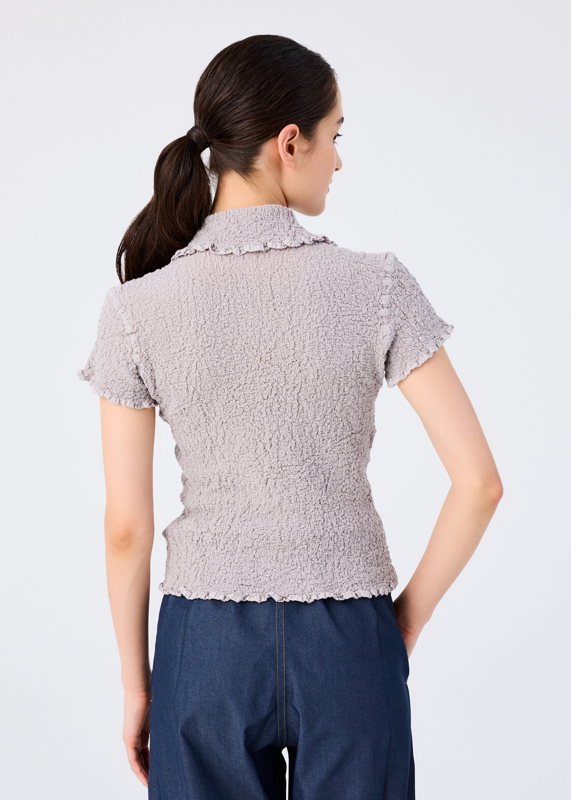 Zoomed view A woman with dark hair in a low ponytail wears the stretchy short-sleeved CAULIFLOWER COLOR top by me ISSEY MIYAKE and dark blue pants, shown from behind against a plain light background.
