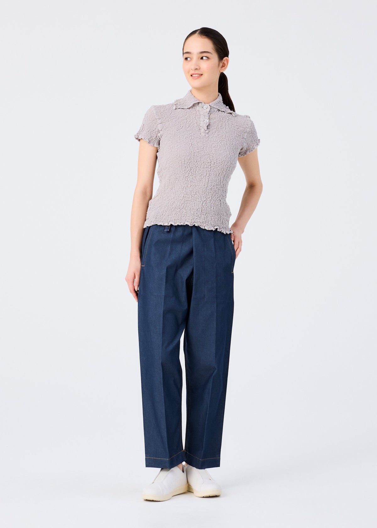 A woman stands against a white background wearing the CAULIFLOWER COLOR top by me ISSEY MIYAKE, paired with wide-leg blue denim pants and white shoes. She looks to the side with one hand behind her back. view 7 of 7