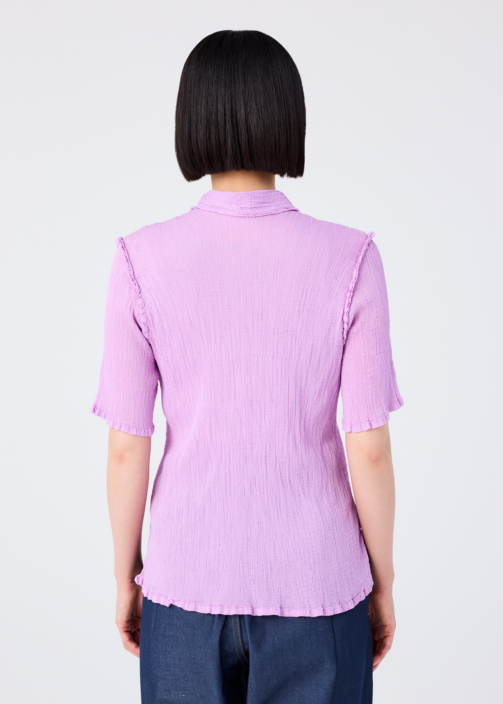 Zoomed view A person with straight, chin-length black hair is seen from behind, wearing the me ISSEY MIYAKE SEE-THROUGH CREPE COLOR light purple collared short-sleeved top and dark blue pants against a plain light background.