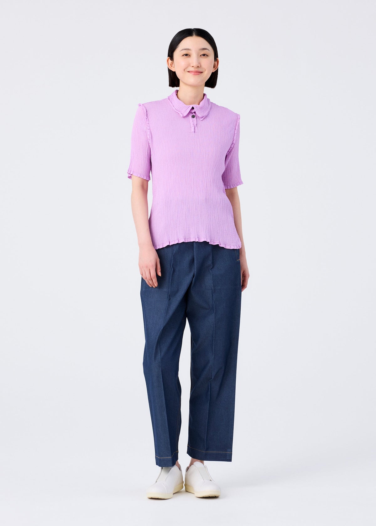 A person stands smiling in a me ISSEY MIYAKE SEE-THROUGH CREPE COLOR light purple collared short-sleeved top, loose blue trousers, and white shoes against a plain white background. view 8 of 9