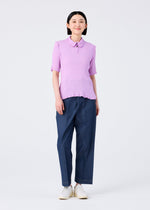 A person stands smiling in a me ISSEY MIYAKE SEE-THROUGH CREPE COLOR light purple collared short-sleeved top, loose blue trousers, and white shoes against a plain white background.