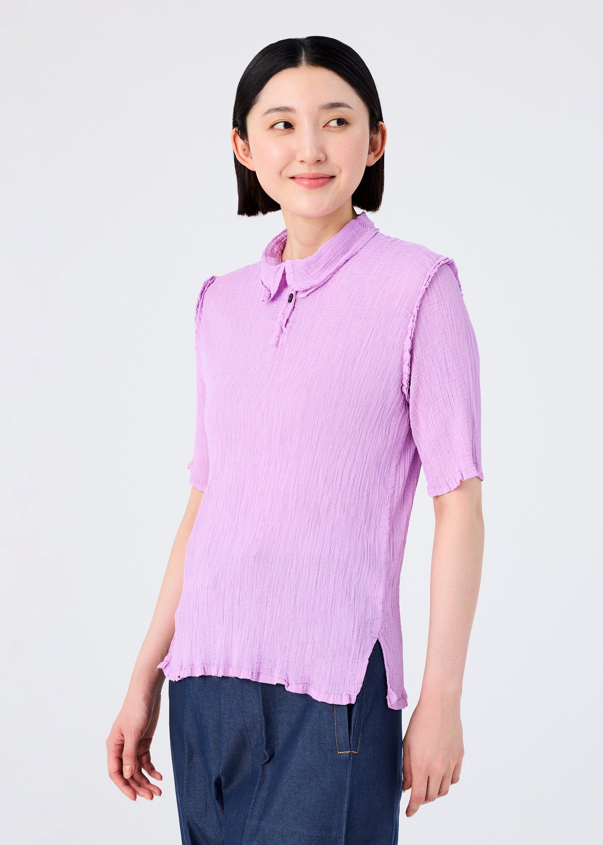 A woman with short dark hair wears the SEE-THROUGH CREPE COLOR blouse by me ISSEY MIYAKE in light purple with short sleeves and textured detail. She stands against a plain white background, looking left and smiling gently. view 9 of 9