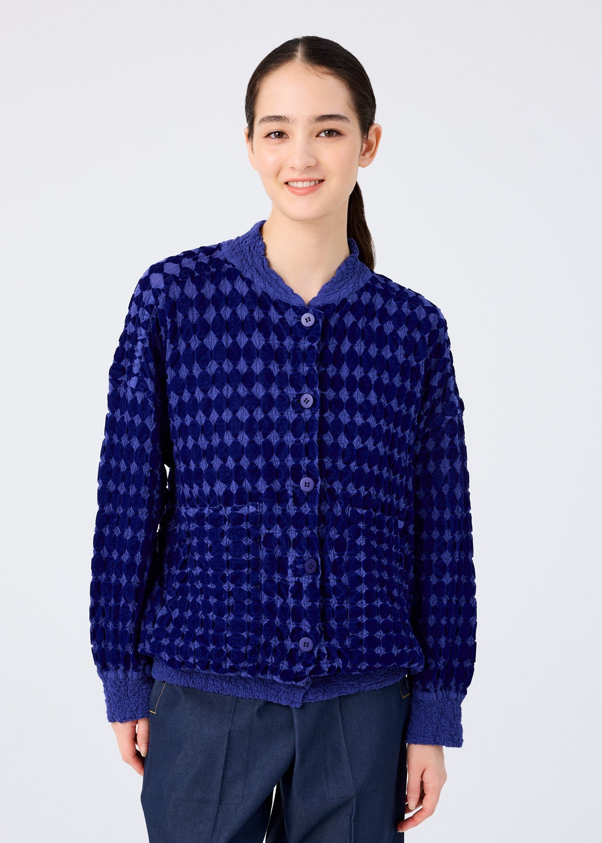 A young woman with long dark hair smiles in the me ISSEY MIYAKE DIAMOND FLOCKY CAULIFLOWER cardigan, featuring a diamond-patterned flock print and long sleeves, paired with dark pants against a plain white background. view 5 of 7