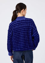 A woman with dark hair in a low ponytail faces away, wearing the me ISSEY MIYAKE DIAMOND FLOCKY CAULIFLOWER long-sleeved cardigan with diamond-patterned flock printing and dark pants against a plain light background.