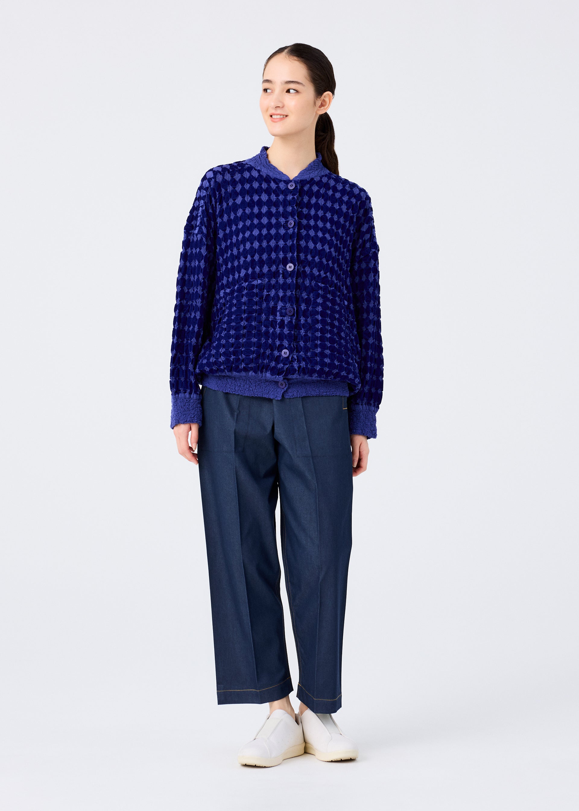 Zoomed view A person stands against a plain white background, wearing the DIAMOND FLOCKY CAULIFLOWER blue cardigan by me ISSEY MIYAKE, loose-fitting dark blue pants, and white shoes. They look slightly to the side and smile.