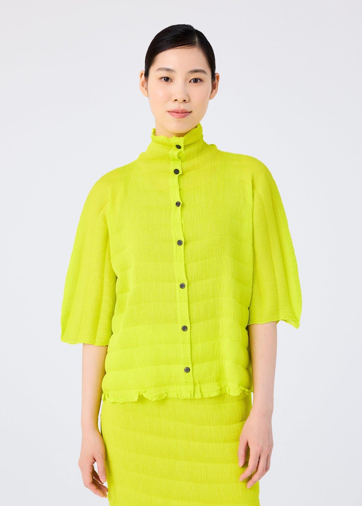 A woman with dark hair tied back models the me ISSEY MIYAKE RIPPLE SEE-THROUGH CREPE, a bright yellow-green pleated blouse with black buttons and matching skirt, against a plain white background. view 5 of 7