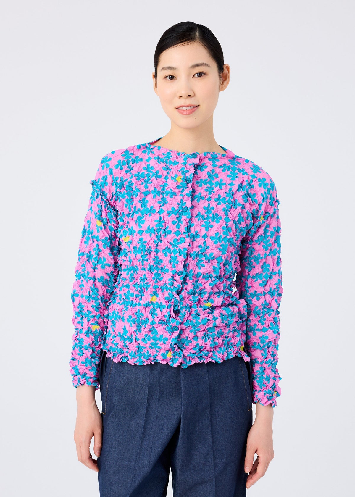 A woman with dark hair pulled back smiles at the camera, wearing the SUIT MERINGUE TOP by me ISSEY MIYAKE—a pink and blue floral long-sleeve blouse with ruffle details—paired with dark blue trousers against a plain background. view 6 of 8