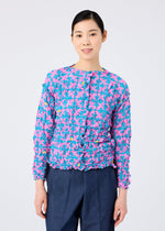 A woman with dark hair pulled back smiles at the camera, wearing the SUIT MERINGUE TOP by me ISSEY MIYAKE—a pink and blue floral long-sleeve blouse with ruffle details—paired with dark blue trousers against a plain background.