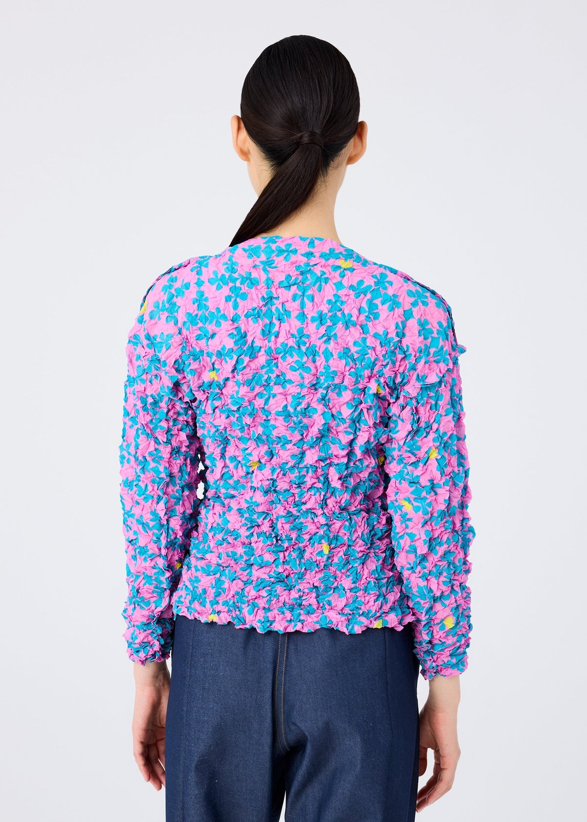 A person with long dark hair in a low ponytail is seen from behind, wearing the SUIT MERINGUE TOP by me ISSEY MIYAKE, featuring a pink, blue, and yellow floral print with subtle card symbols, paired with dark blue pants against a plain background. view 7 of 8