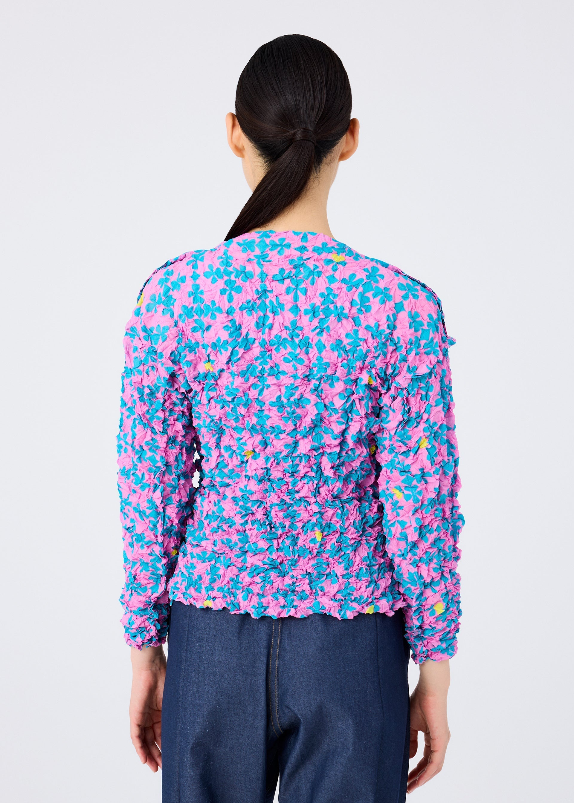 Zoomed view A person with long dark hair in a low ponytail is seen from behind, wearing the SUIT MERINGUE TOP by me ISSEY MIYAKE, featuring a pink, blue, and yellow floral print with subtle card symbols, paired with dark blue pants against a plain background.