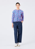 A person with short dark hair and a neutral expression stands against a white background, wearing the SUIT MERINGUE TOP by me ISSEY MIYAKE in textured blue and pink, loose blue trousers, and white shoes.