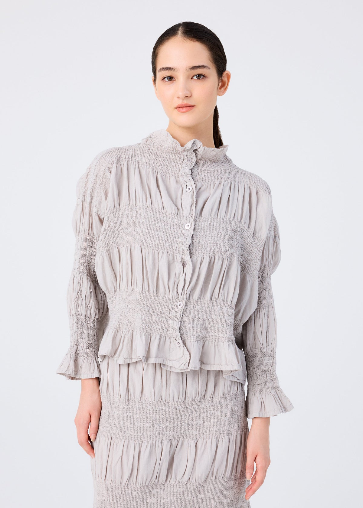 A woman with long dark hair in a low ponytail wears the SHRINK STRIPE blouse and skirt set by me ISSEY MIYAKE, featuring light gray pleated textured fabric, standing against a plain white background. view 5 of 7