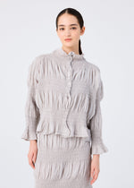 A woman with long dark hair in a low ponytail wears the SHRINK STRIPE blouse and skirt set by me ISSEY MIYAKE, featuring light gray pleated textured fabric, standing against a plain white background.