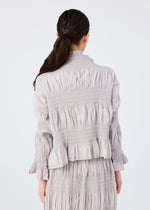 A woman with long dark hair in a low ponytail is seen from behind, wearing the SHRINK STRIPE long-sleeved top and matching skirt by me ISSEY MIYAKE, both featuring gathered and ruched details, against a plain background.