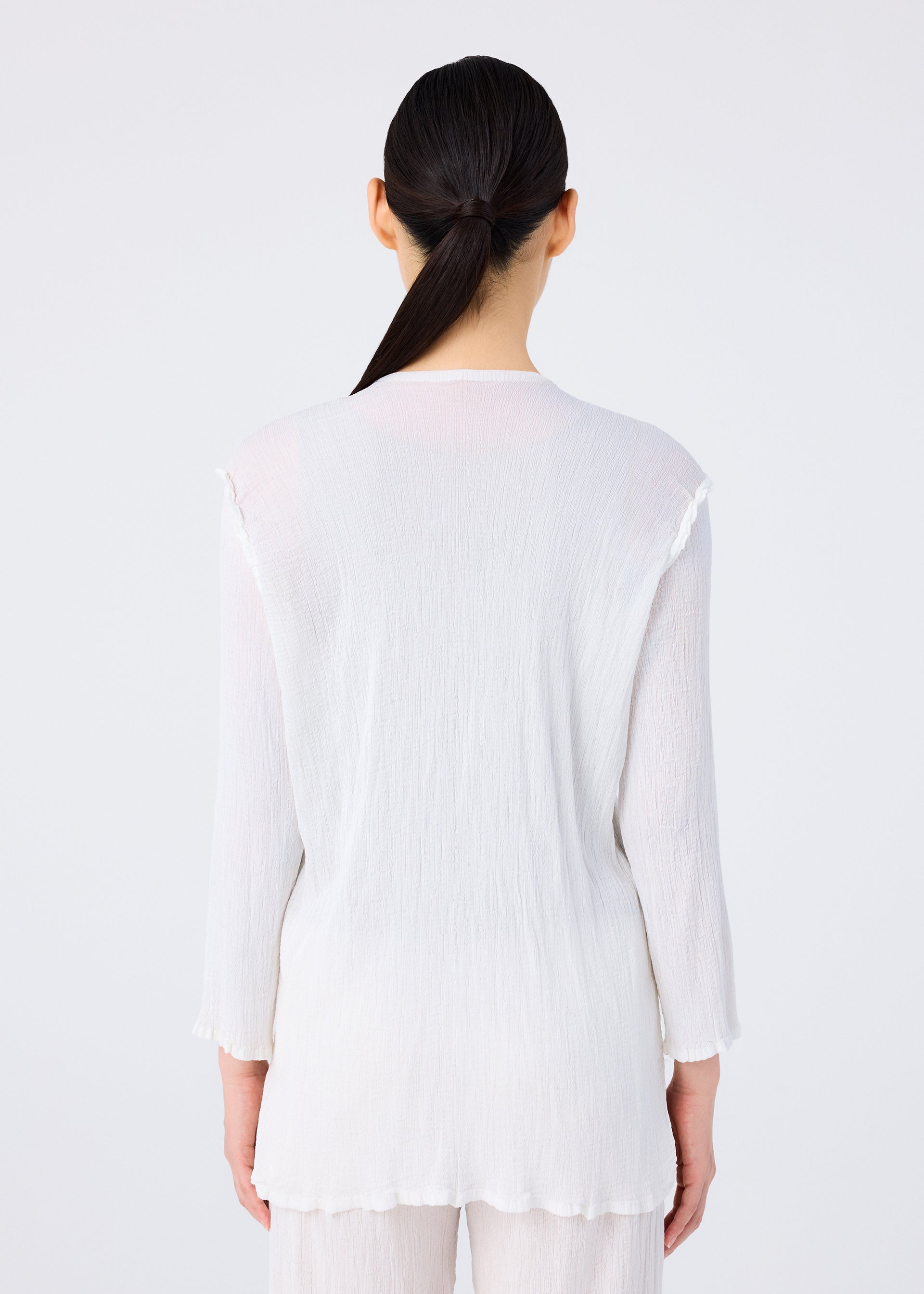 Zoomed view A woman with long dark hair in a low ponytail stands with her back to the camera, wearing the SEE-THROUGH CREPE COLOR top and matching pants by me ISSEY MIYAKE, against a plain light background.