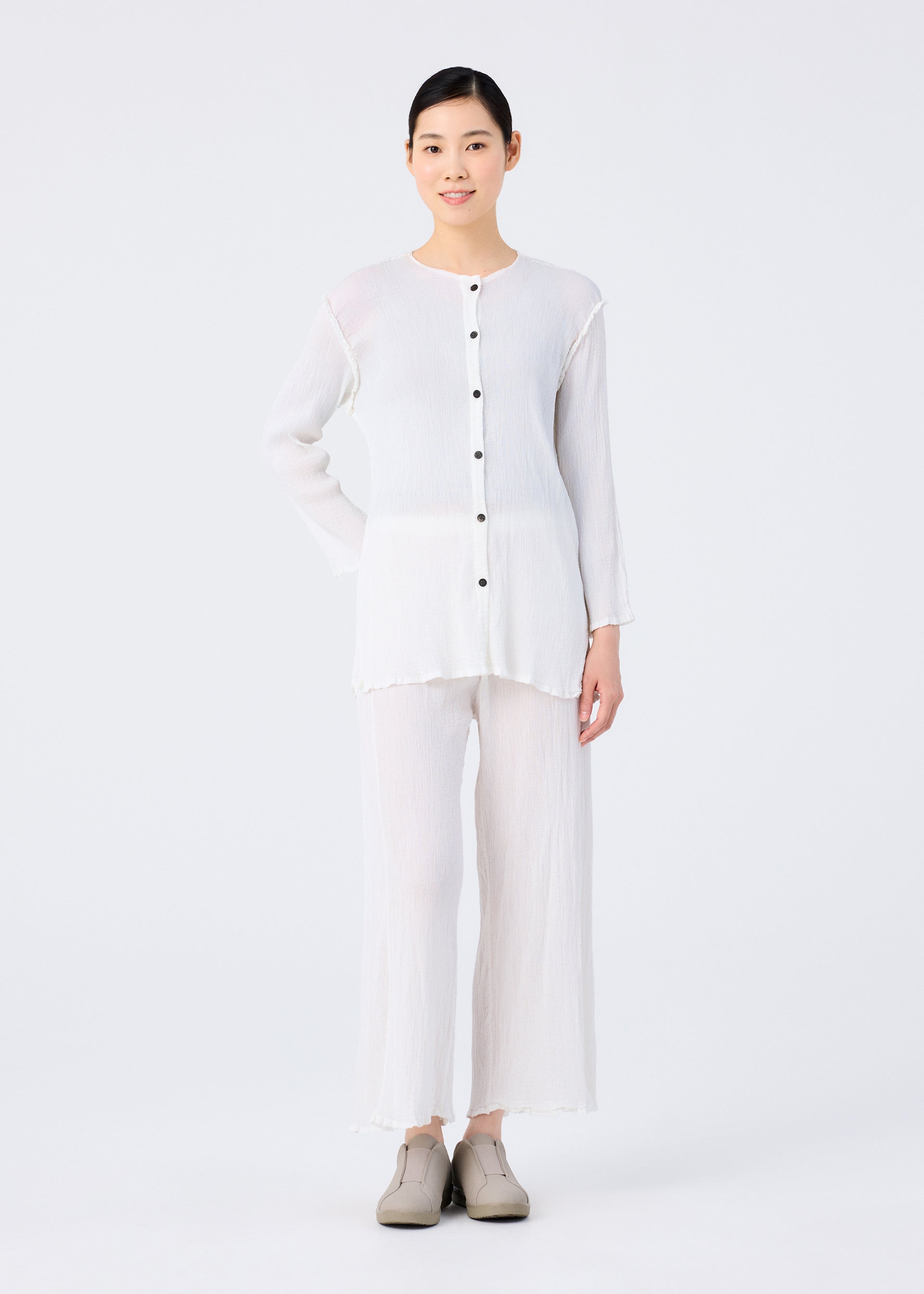Zoomed view A person with short dark hair wears the me ISSEY MIYAKE SEE-THROUGH CREPE COLOR cardigan—loose-fitting, long-sleeved, garment-dyed with buttons—matched with wide-legged white pants and beige shoes against a plain white background.