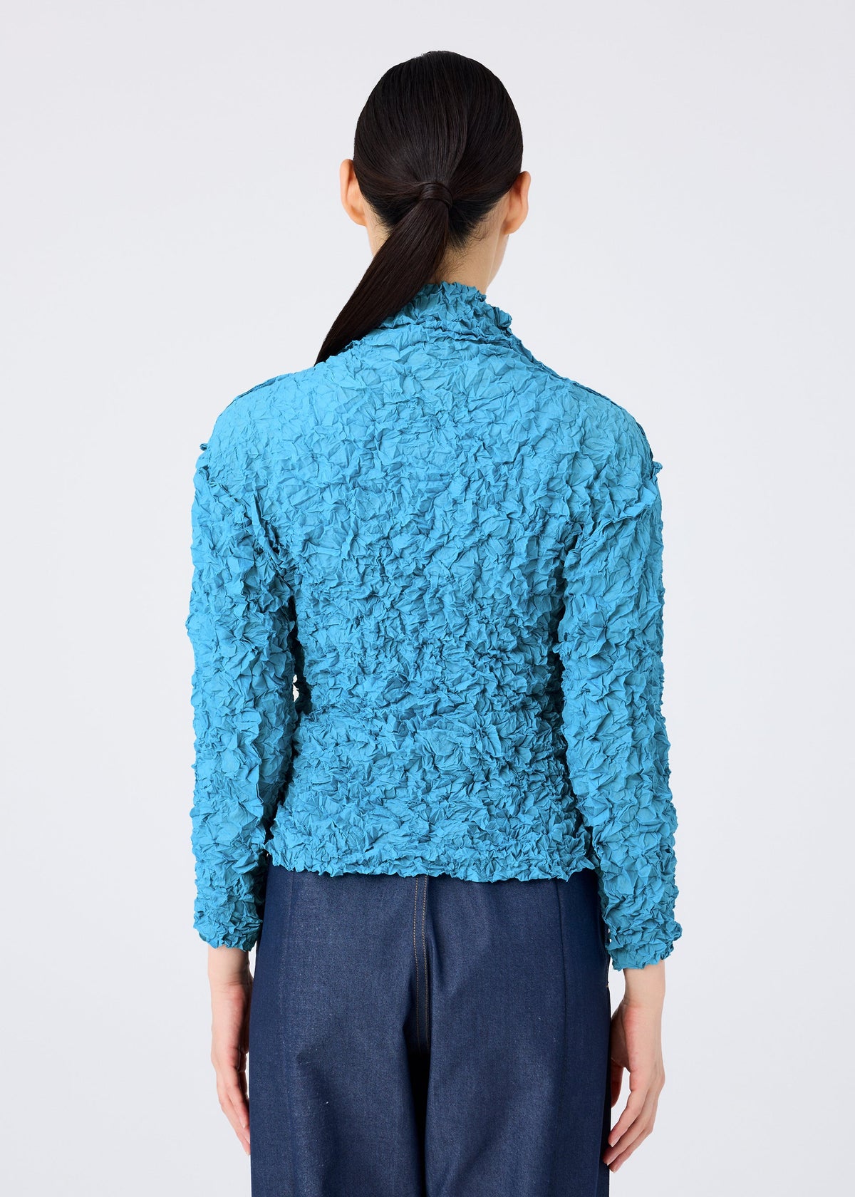 Seen from behind, a person with long dark hair in a low ponytail wears the MERINGUE TOP by me ISSEY MIYAKE—a textured, bright blue crushed pleat long-sleeve top—with dark blue pants against a plain white background. view 9 of 10