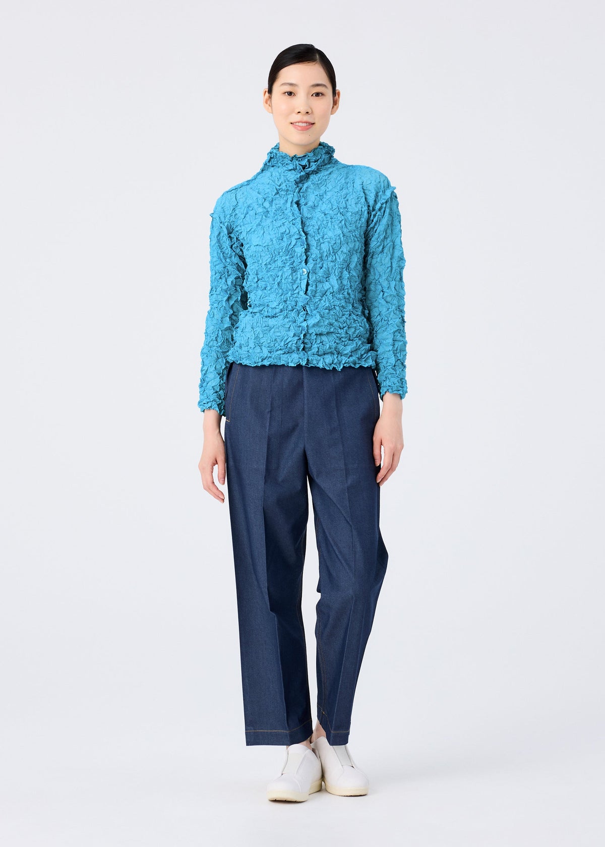 A woman stands facing forward against a plain white background, wearing the MERINGUE TOP by me ISSEY MIYAKE in textured bright blue, paired with loose dark blue trousers and white shoes. view 10 of 10