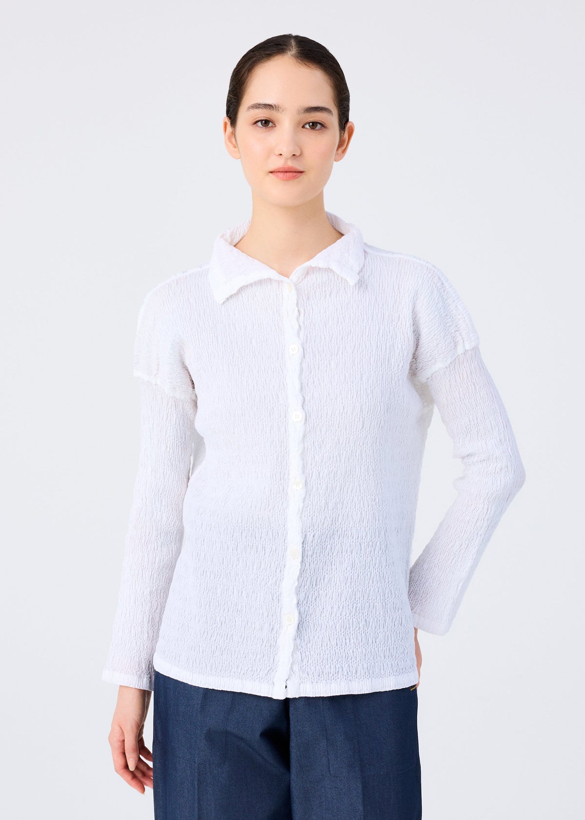 A woman with dark hair tied back models the me ISSEY MIYAKE KNIT LIKE CLOTH textured white cotton blend button-up long-sleeve shirt and dark blue pants, ideal for spring and summer, against a plain light background. view 5 of 7