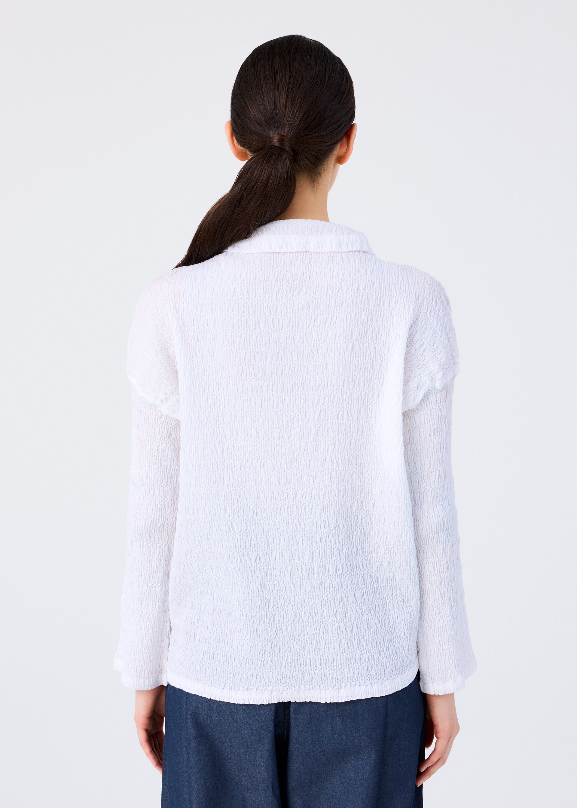Zoomed view A woman with long dark hair in a low ponytail is seen from behind, wearing me ISSEY MIYAKE’s KNIT LIKE CLOTH white cotton-blend cardigan and dark blue pants—ideal for spring and summer—against a simple light background.