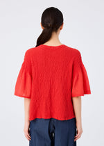 A person with long dark hair in a low ponytail is seen from behind, wearing the SHEER CAULIFLOWER cardigan by me ISSEY MIYAKE in red, paired with dark blue pants, set against a plain light background.