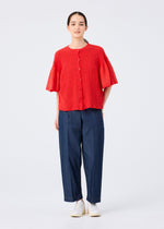 A person stands against a plain background wearing the SHEER CAULIFLOWER red cotton short-sleeved cardigan by me ISSEY MIYAKE, wide-leg blue pants, and white shoes. Their hair is tied back, facing forward with a neutral expression.