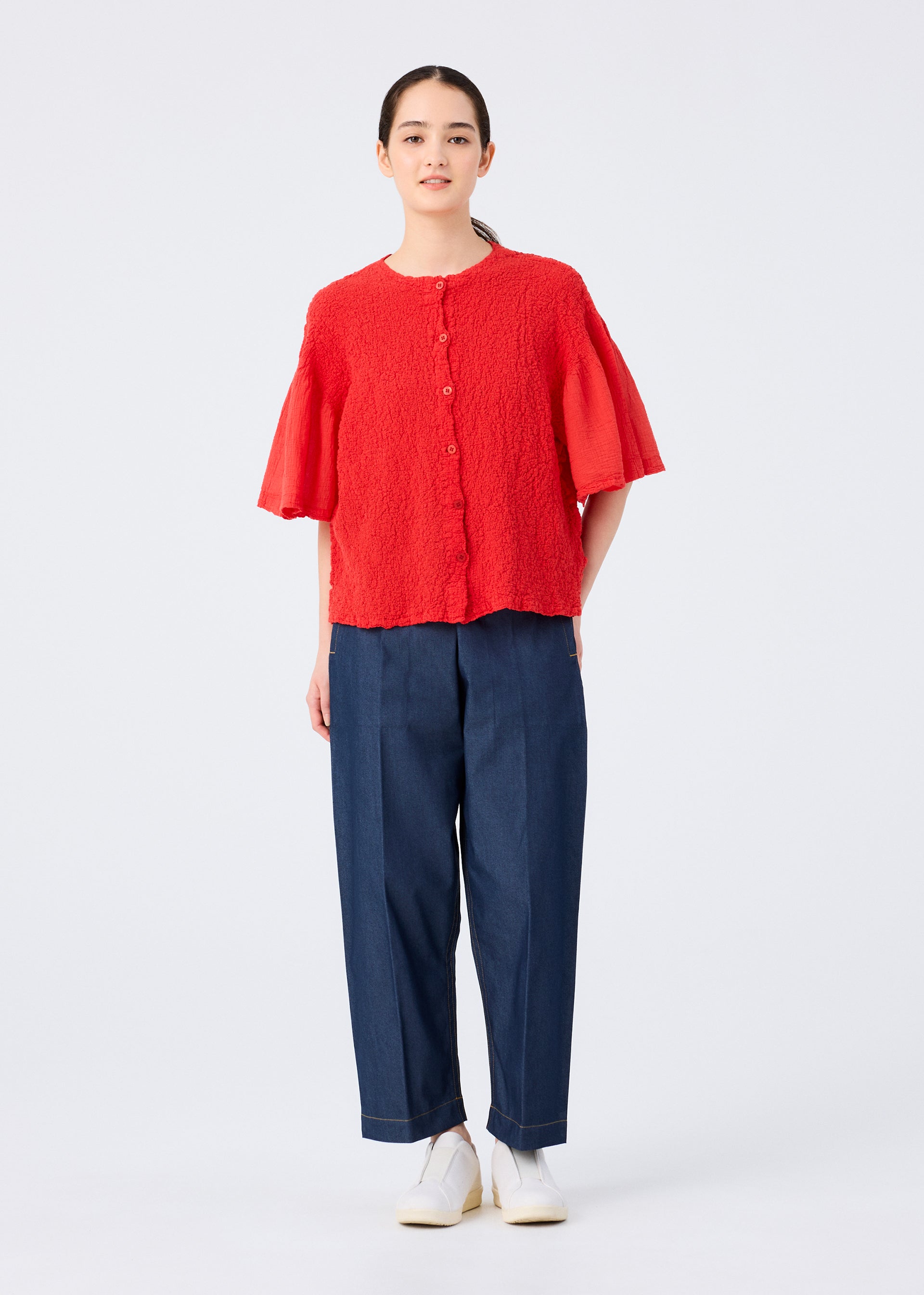 Zoomed view A person stands against a plain background wearing the SHEER CAULIFLOWER red cotton short-sleeved cardigan by me ISSEY MIYAKE, wide-leg blue pants, and white shoes. Their hair is tied back, facing forward with a neutral expression.