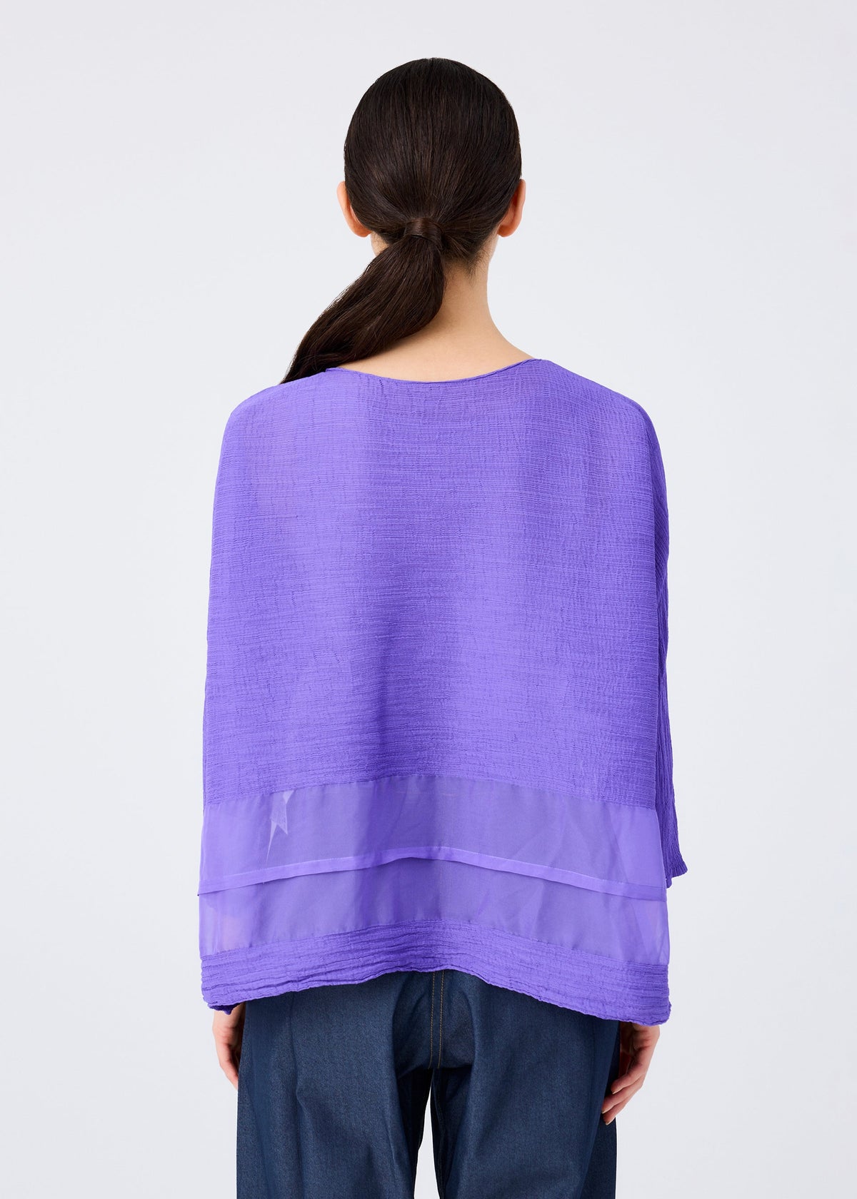 A person with long dark hair in a low ponytail is shown from behind, wearing the me ISSEY MIYAKE SHEER SEE-THROUGH CREPE top in textured purple with a sheer band and dark blue pants, set against a plain white background. view 6 of 7