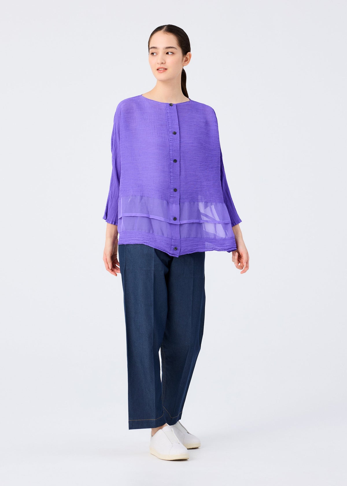 A woman with long dark hair tied back models the me ISSEY MIYAKE SHEER SEE-THROUGH CREPE, a loose-fitting, garment-dyed purple button-up top with sheer layers, paired with wide-leg dark blue jeans and white shoes. view 7 of 7