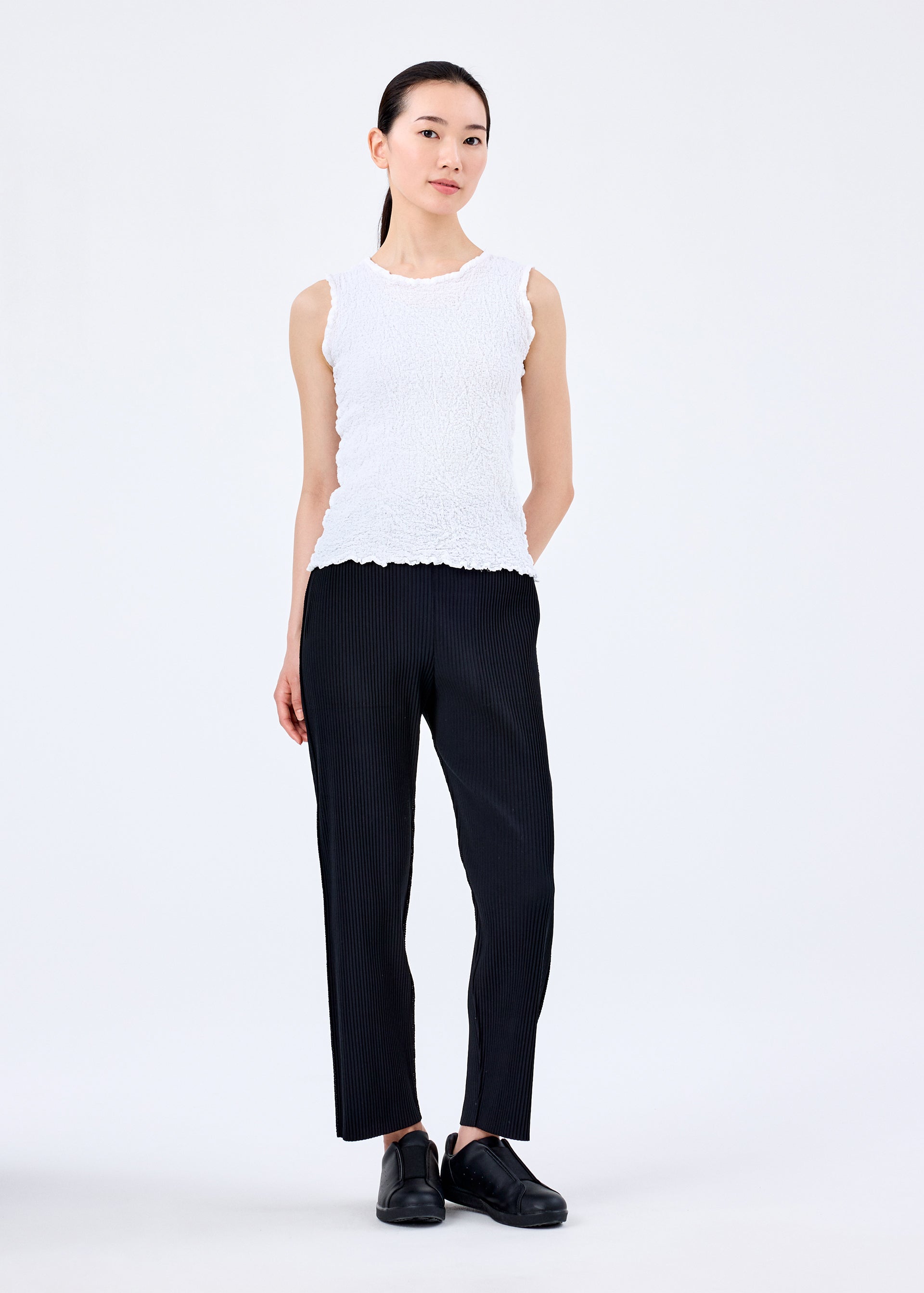 Zoomed view A woman with a neutral expression stands against a white background, wearing the CAULIFLOWER sleeveless cotton round-neck top by me ISSEY MIYAKE, paired with black pinstripe pants and shoes, her left hand behind her back.